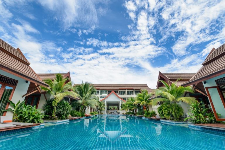 Hồ bơi dài giữa các toà nhà mái dốc và hàng cọ của một resort nhiệt đới dưới trời xanh