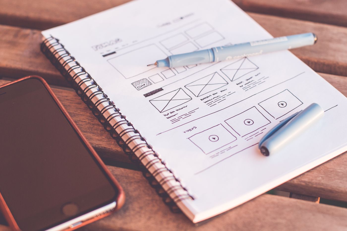 Sổ tay gáy lò xo với bản phác thảo khung giao diện website, bên cạnh là bút và điện thoại trên bàn gỗ