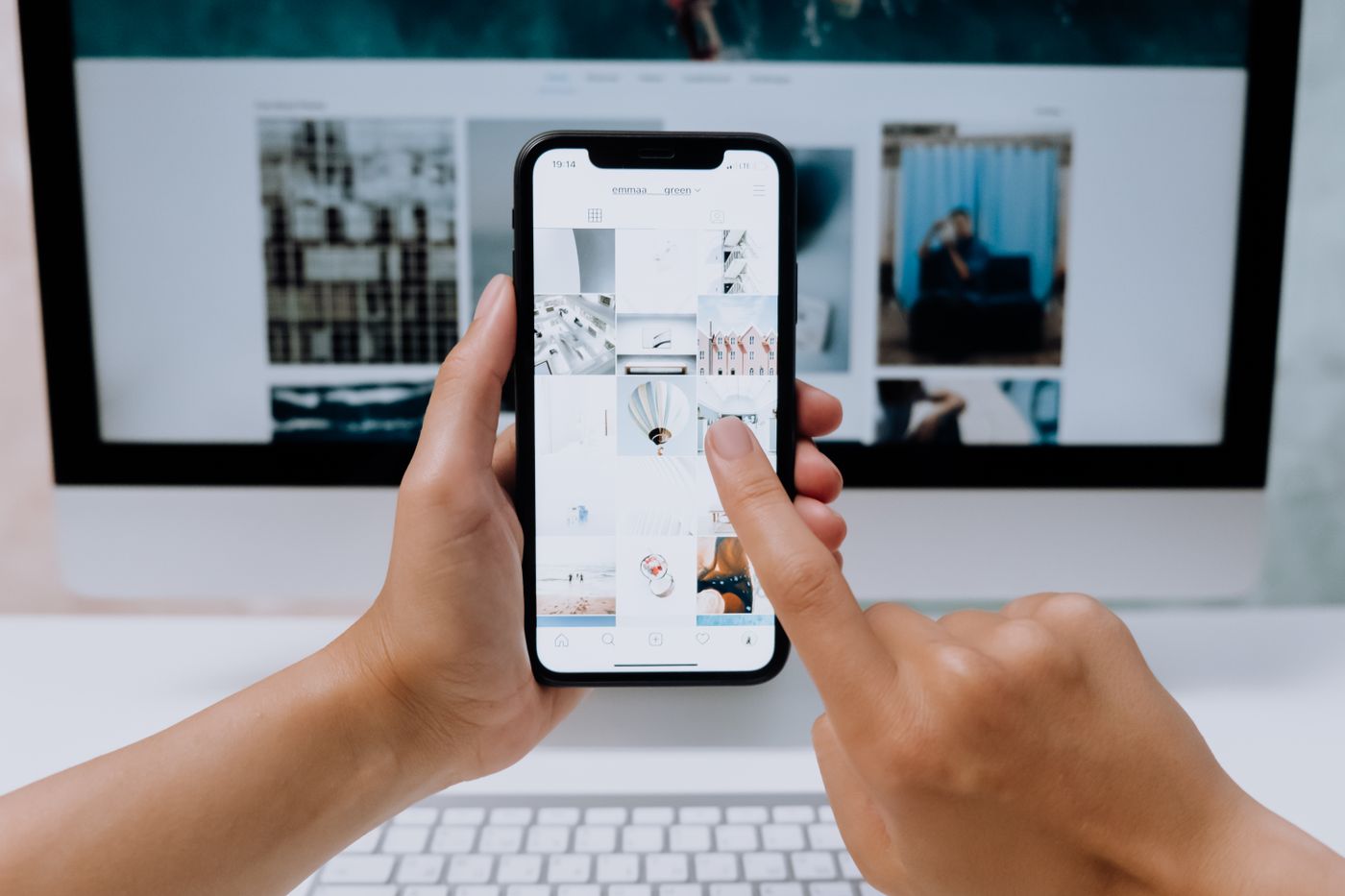Bàn tay cầm điện thoại xem một trang web, phía sau là màn hình máy tính hiển thị cùng trang web
