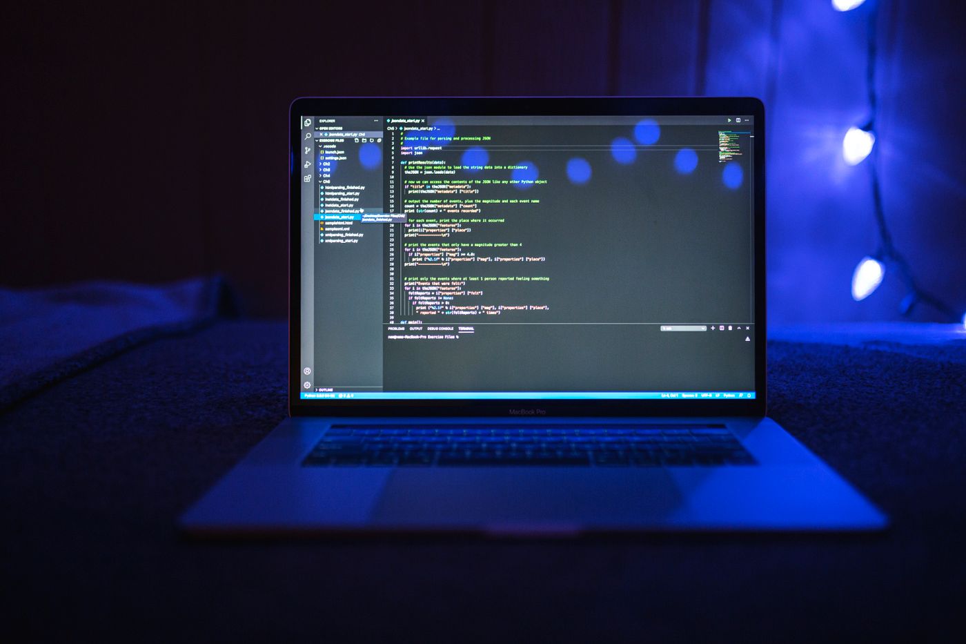 Laptop đặt trên bàn trong phòng tối, màn hình hiển thị mã nguồn, phía sau là ánh đèn xanh dương