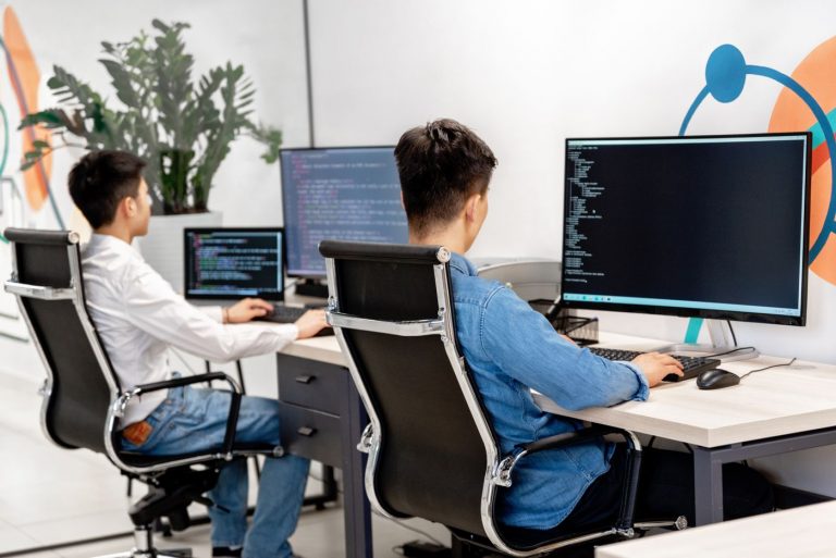 Hai lập trình viên ngồi ghế lưới đen làm việc trước các màn hình hiển thị mã nguồn trong văn phòng sáng