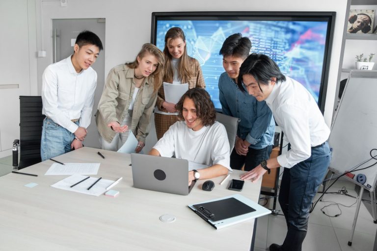 Nhóm đồng nghiệp đứng quanh một chiếc laptop trong phòng họp, cùng xem màn hình